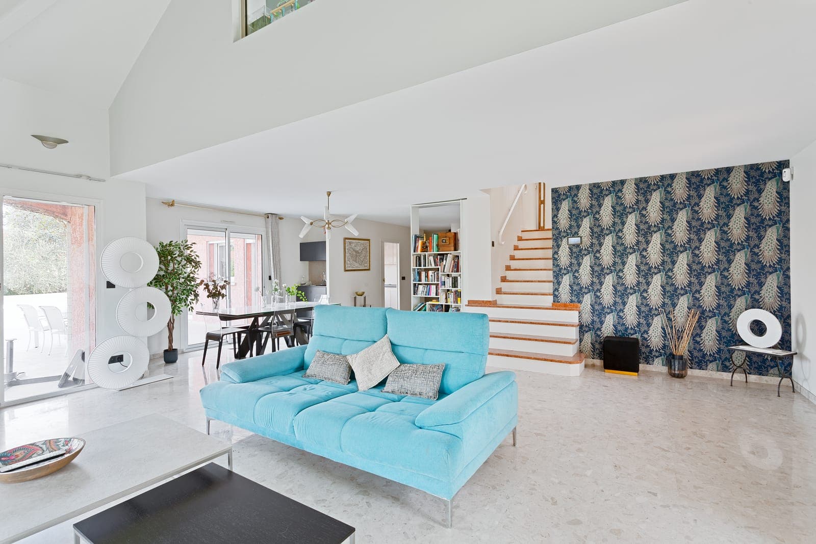 Séjour cathédrale blanc et lumineux avec canapé turquoise, escalier et papier peint décoratif, dans une villa rénovée à Bron
