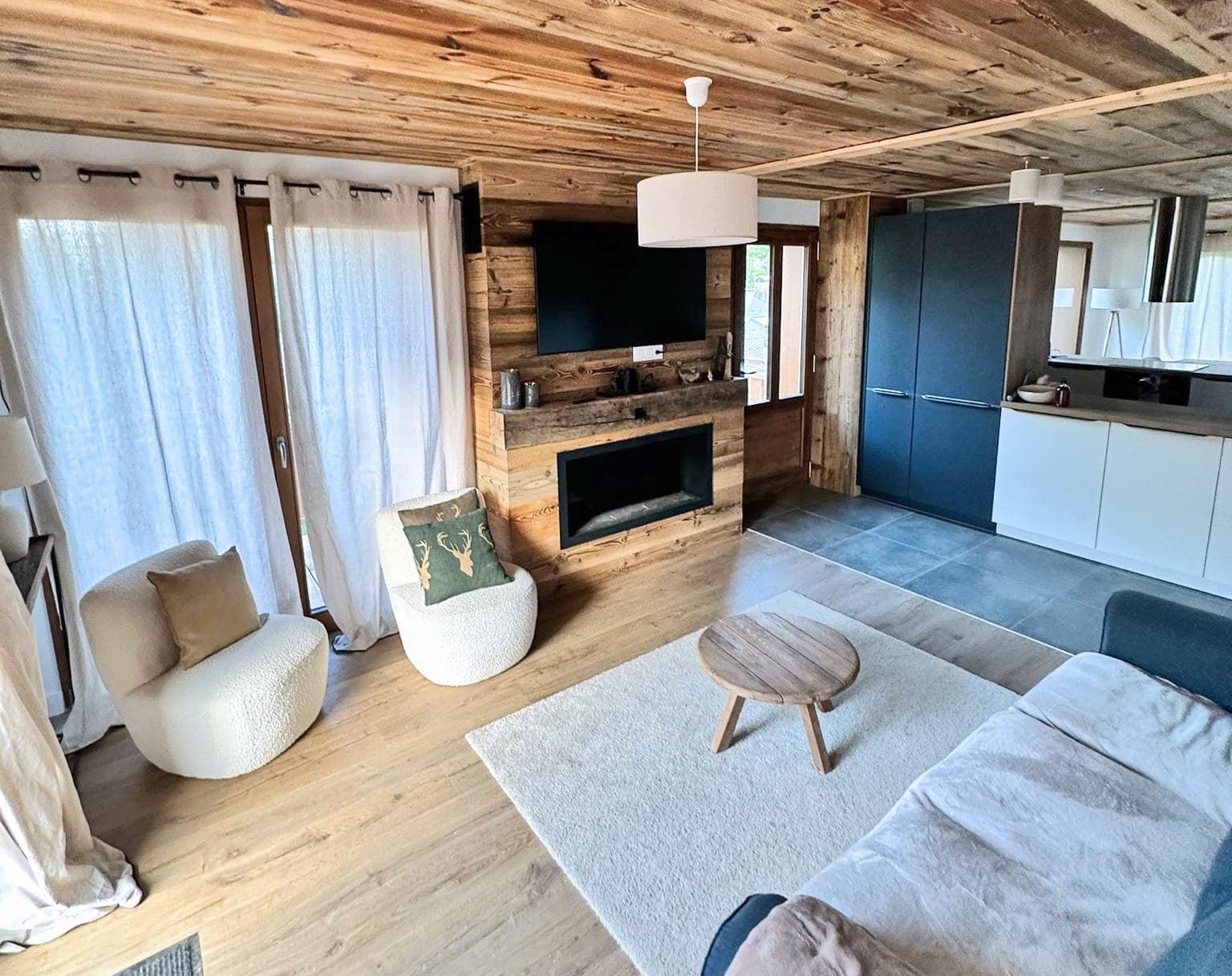 Salon de chalet avec cheminée en bois vieilli, canapé clair et cuisine bleu marine, au chalet Le Nard Bleu à Praz-sur-Arly