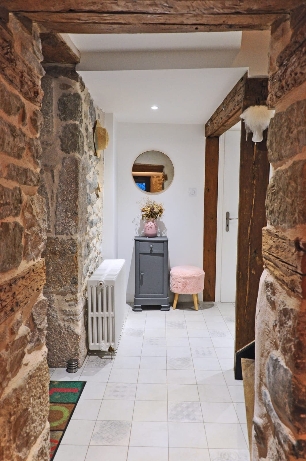 Entrée aux murs de pierre et poutres anciennes, avec miroir rond et petit meuble gris, dans une maison rénovée à Sauverny