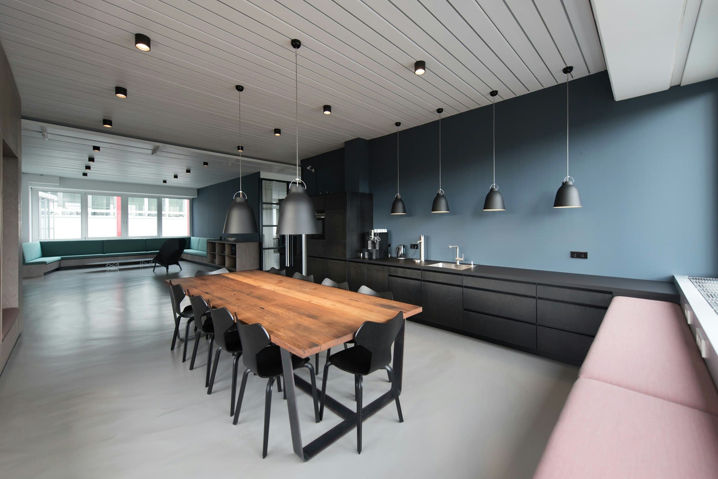 Cafétéria de bureaux, grande table en bois sous des suspensions et cuisine équipée