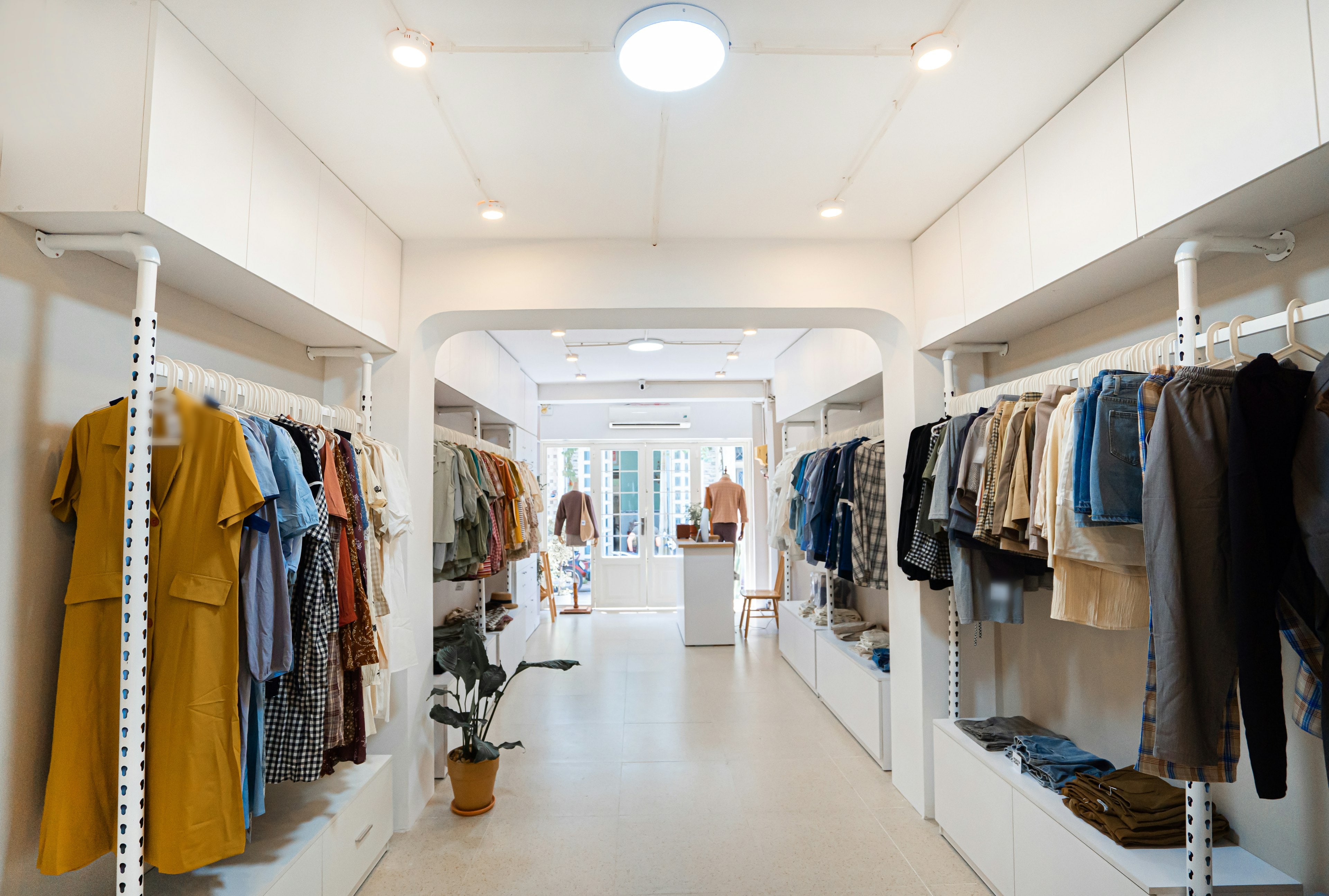 Boutique de prêt-à-porter aux murs blancs, portants de part et d'autre d'une allée menant à une arche lumineuse
