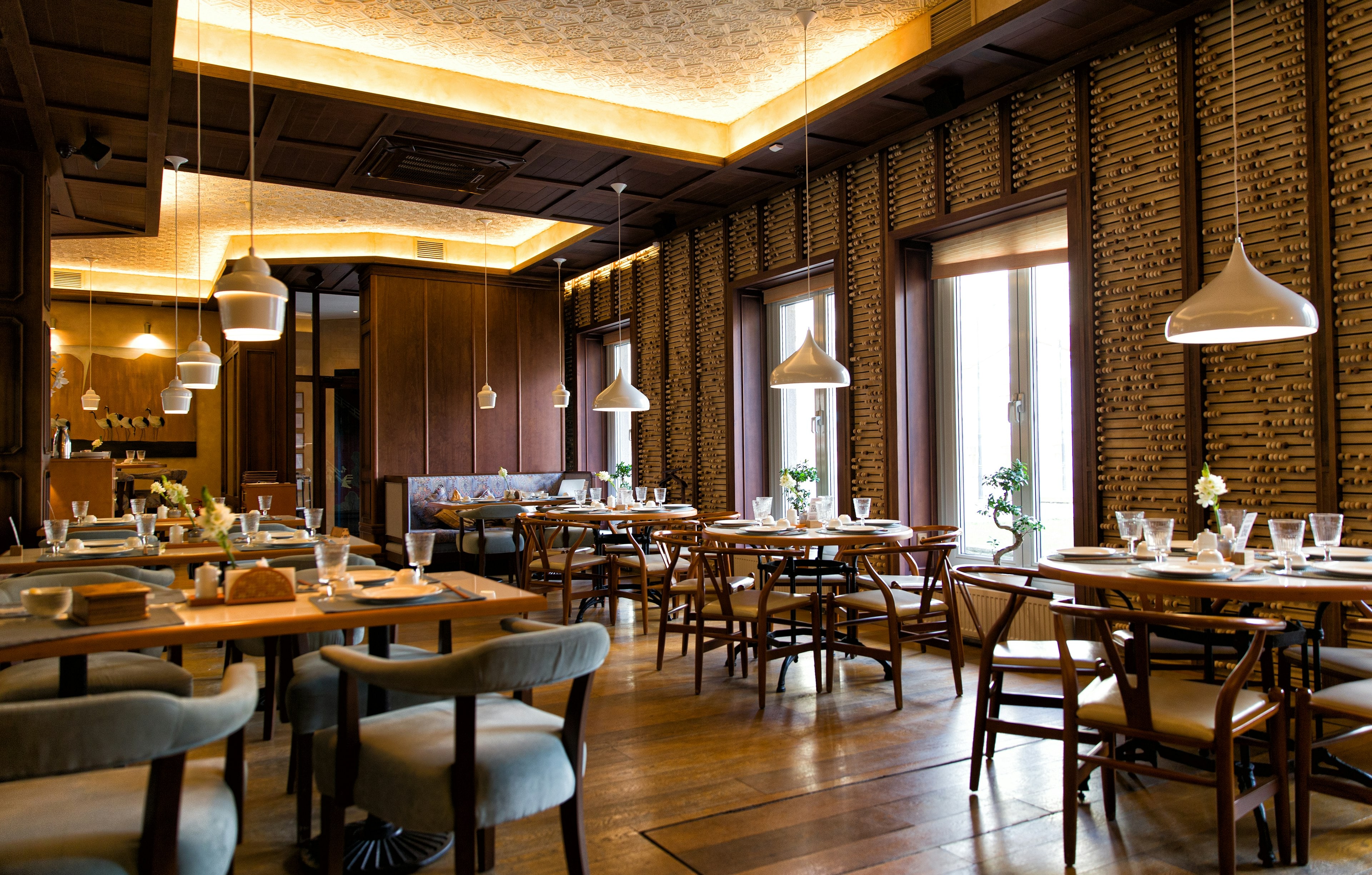Salle de restaurant chaleureuse aux boiseries sombres, suspensions blanches et tables dressées devant de hautes fenêtres