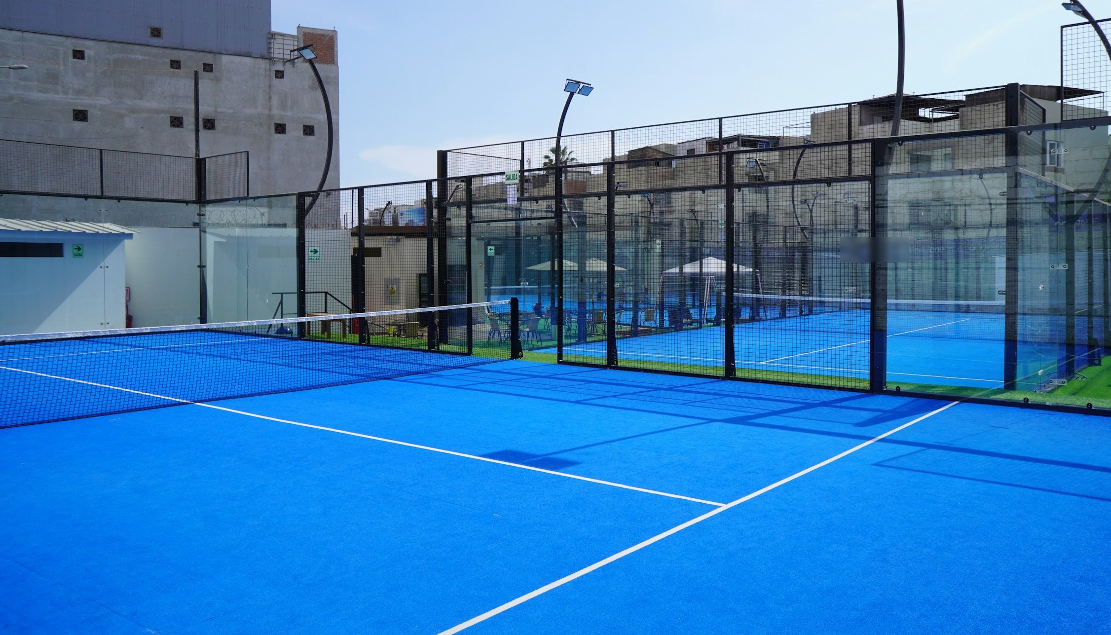 Piste de padel bleue entourée de vitrages, éclairage sur mâts courbes