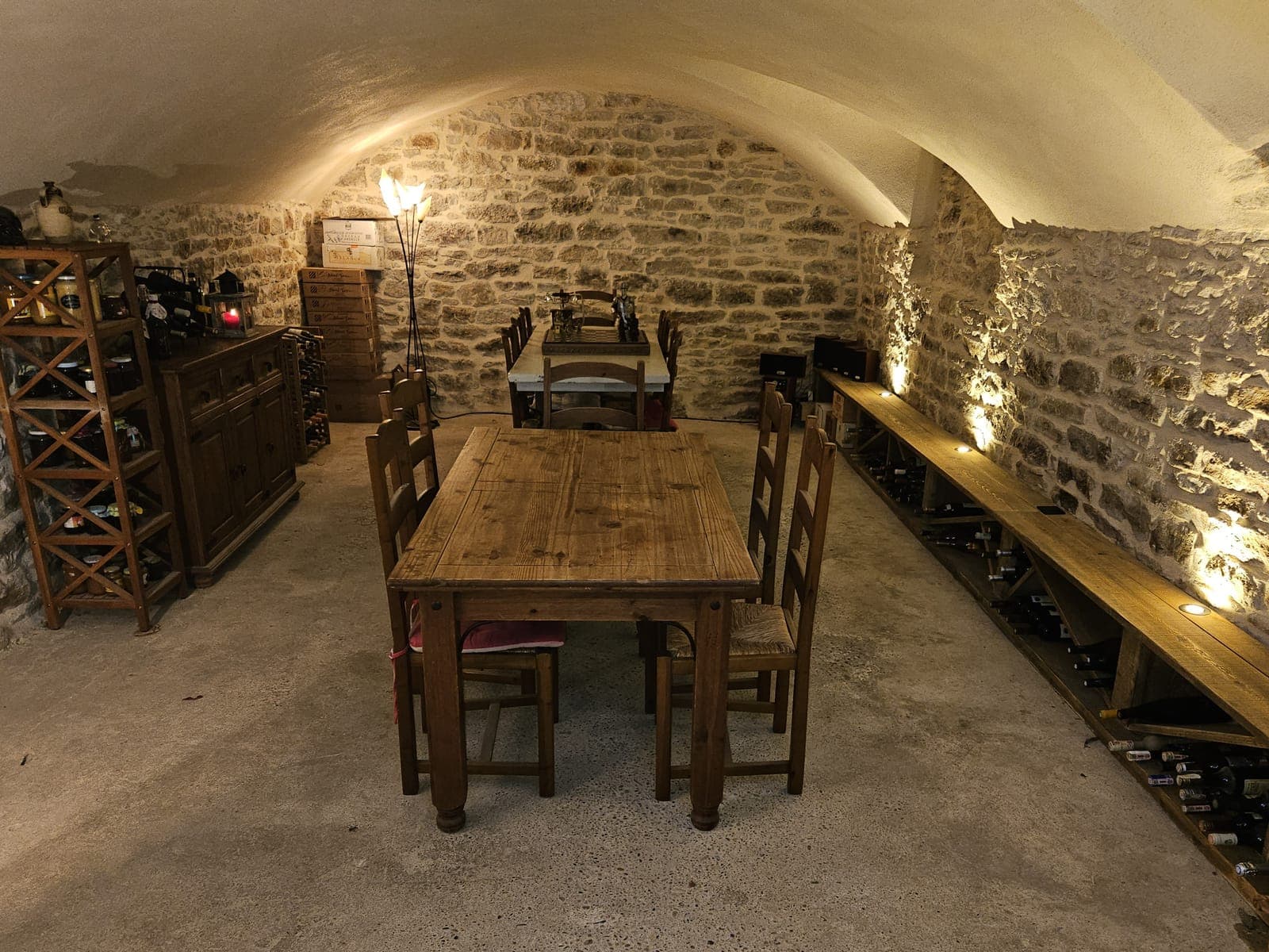 Cave voûtée en pierre éclairée, aménagée avec table en bois et rangements à bouteilles, dans une maison de village à Saint-Vulbas